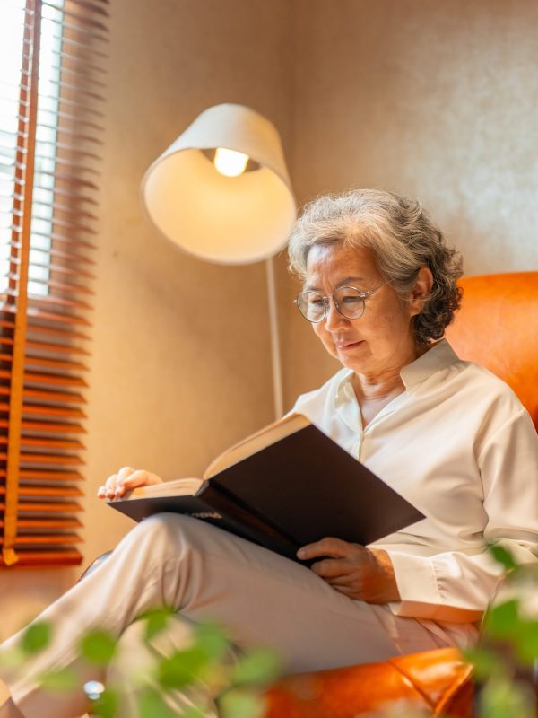 Wellness, mental healthcare in elderly people at home. Happy Asian senior mature woman relaxing on sofa in living room and reading a book. Retired woman enjoy leisure activity hobbies at nursing home.