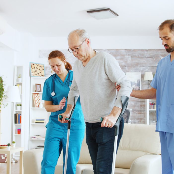 Team of nurses or social workers helping an old disabled man to walk with his crutches out of the nursing home room.