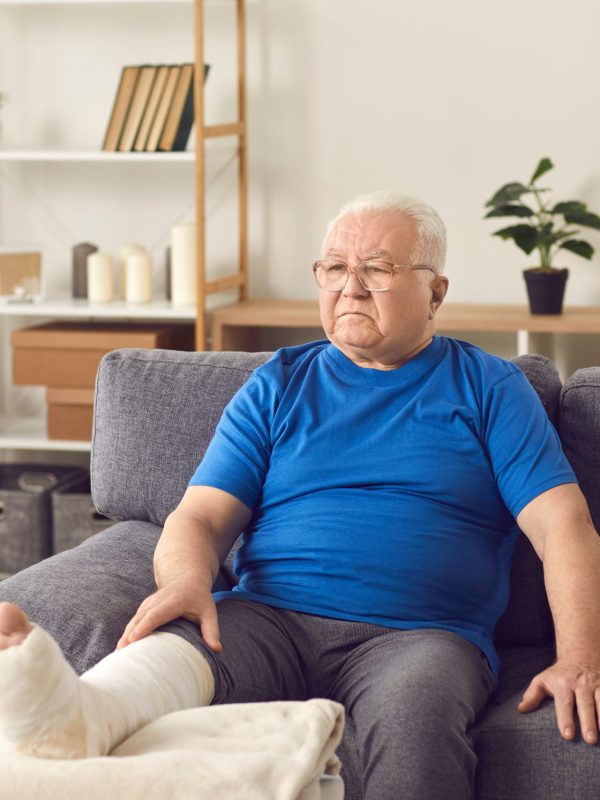Unhappy disappointed senior mature man sitting on sofa with broken leg in cast and crutches nearby and looking aside with room interior at background. Injury, trauma, recovery, rehabilitation concept