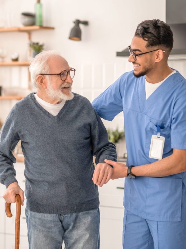 Smiling friendly in-home male nurse in uniform supporting an old man with a walking stick