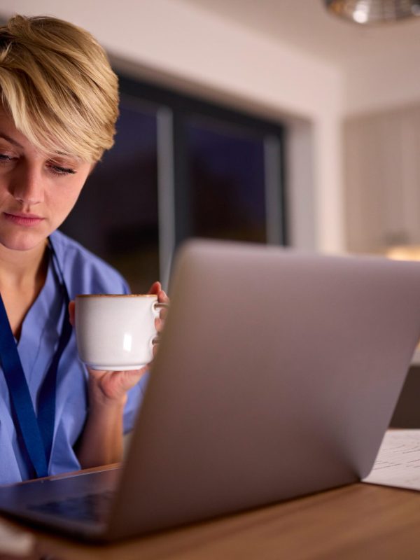 Tired Woman Wearing Medical Scrubs Working Or Studying On Laptop At Home At Night